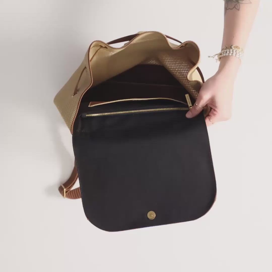 The Cinch Backpack in Brown Butter being packed with a camera, a water bottle, a tube of hand cream, a bottle of sunscreen, a book and a baseball cap. The model then unzips the interior pocket and puts a Wallet wristlet inside, along with an iPhone and a pair of sunglasses. The model cinches the drawstring closed and carries the bag out of frame. 